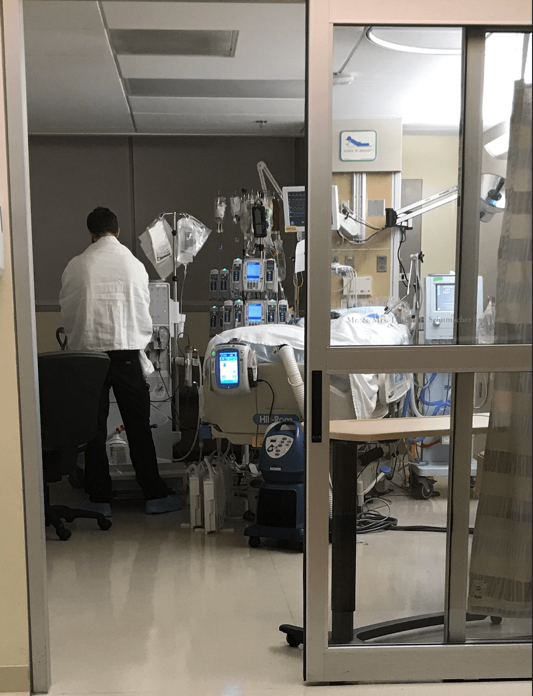 View through a sliding glass ICU door during a COVID travel assignment: a nurse stands at the bedside with IV pumps, monitors, ventilator equipment, and a Hill-Rom bed surrounding the patient.